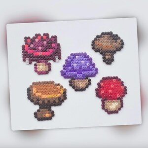 Op de afbeelding: Vijf verschillende paddenstoelontwerpen gemaakt met kleurrijke perler kralen. De paddenstoelen zijn in verschillende kleuren, waaronder rood, paars, bruin en wit.