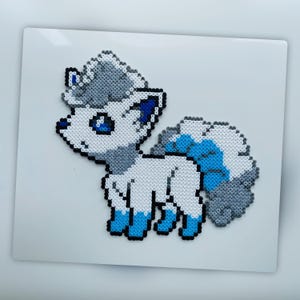 May include: A pixel art depiction of a white and blue fox-like creature. The artwork is made of small, colorful beads, with a fluffy tail and ears. The creature has blue eyes and accents, set against a white background.