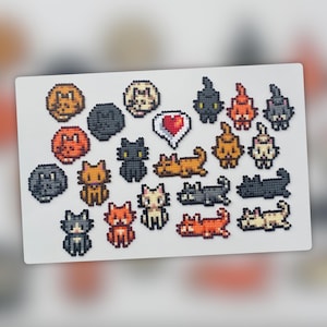 Op de afbeelding: Een verzameling gepixelde katten- en hondenontwerpen in verschillende kleuren, waaronder oranje, zwart, grijs en wit. De ontwerpen zijn gerangschikt op een witte achtergrond, met een gepixeld hart in het midden. Het thema is een schattig, handgemaakt ambacht.