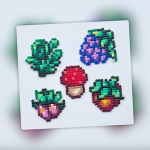 Op de afbeelding: Vijf kleurrijke pixel art-ontwerpen gemaakt met kralen. De ontwerpen omvatten een ​​rode paddenstoel, een paarse bloem met groene bladeren, een groene plant en twee rode aardbeien met groene bladeren.