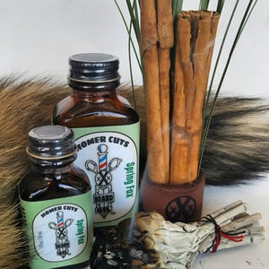 May include: Two amber glass bottles with green labels that read "Homer Cuts Spring Fox Beard Oil" and a bundle of cinnamon sticks in a leather holder. A smoldering white sage bundle is in the foreground.