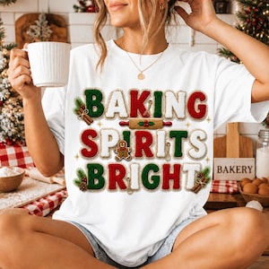 May include: A white t-shirt featuring the text BAKING SPIRITS BRIGHT in red and green textured letters. The design includes a gingerbread man, a rolling pin, and pine sprigs. The letters have a stitched border effect, creating a festive look.