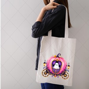 Puede incluir: Un bolso tote blanco con un dise&ntilde;o de carruaje de calabaza rosa y un fantasma dentro. El carruaje tiene detalles dorados, cortinas moradas y ruedas negras. Dos faroles flanquean el carruaje, ideal para Halloween.