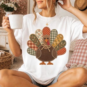 Puede incluir: Una camiseta blanca de manga corta con un gráfico central de un pavo compuesto por varios parches de tela estampada. El diseño del pavo incluye texturas de cuadros, flores y lunares en tonos marrones, naranjas, verdes y crema.