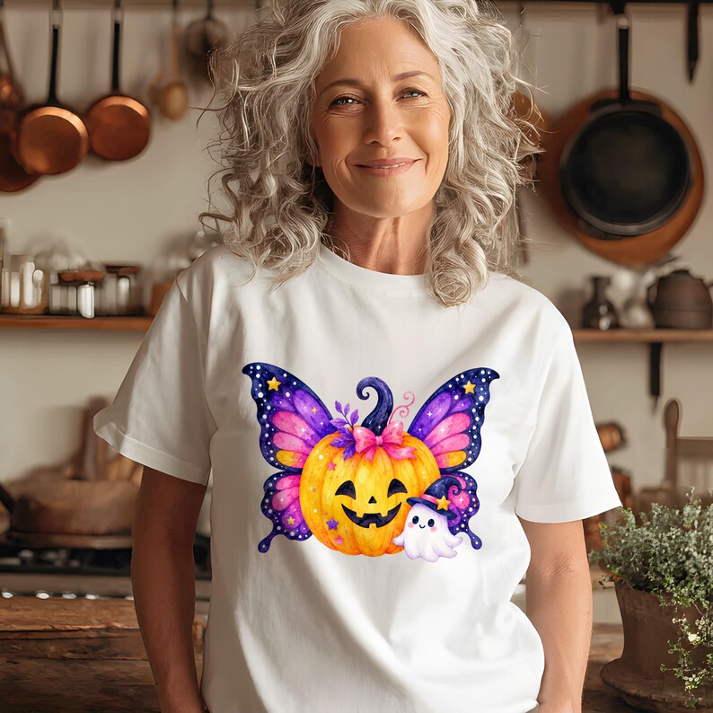 Puede incluir: Camiseta blanca con un dise&ntilde;o tem&aacute;tico de Halloween. El gr&aacute;fico incluye una calabaza sonriente con alas de mariposa en tonos morados, rosas y amarillos, un peque&ntilde;o fantasma y un sombrero de bruja.
