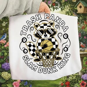 May include: White graphic tee featuring a raccoon wearing sunglasses and a checkered pattern. The raccoon is holding a basketball and the text reads "Trash Panda Slam Dunk King" in a circular design.
