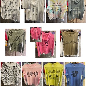 Op de afbeelding: Collectie shirts met lange mouwen en hoodies met diverse grafische ontwerpen. De shirts zijn in kleuren als grijs, roze, geel en blauw. Ontwerpen omvatten gekko's, slangen en plantmotieven. Sommige shirts hebben de tekst "Geckos of New Caledonia".