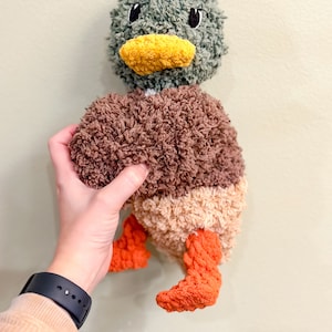 Op de afbeelding: Een pluche eendenspeelgoed met een bruin, beige en oranje lichaam. De eend heeft een groene kop met een gele snavel en zwarte ogen.