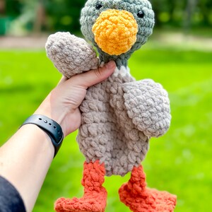 Op de afbeelding: Een gehaakt pluche speelgoed eend met een grijs lichaam, een groene kop, een oranje snavel en oranje poten. De eend wordt vastgehouden door een hand.