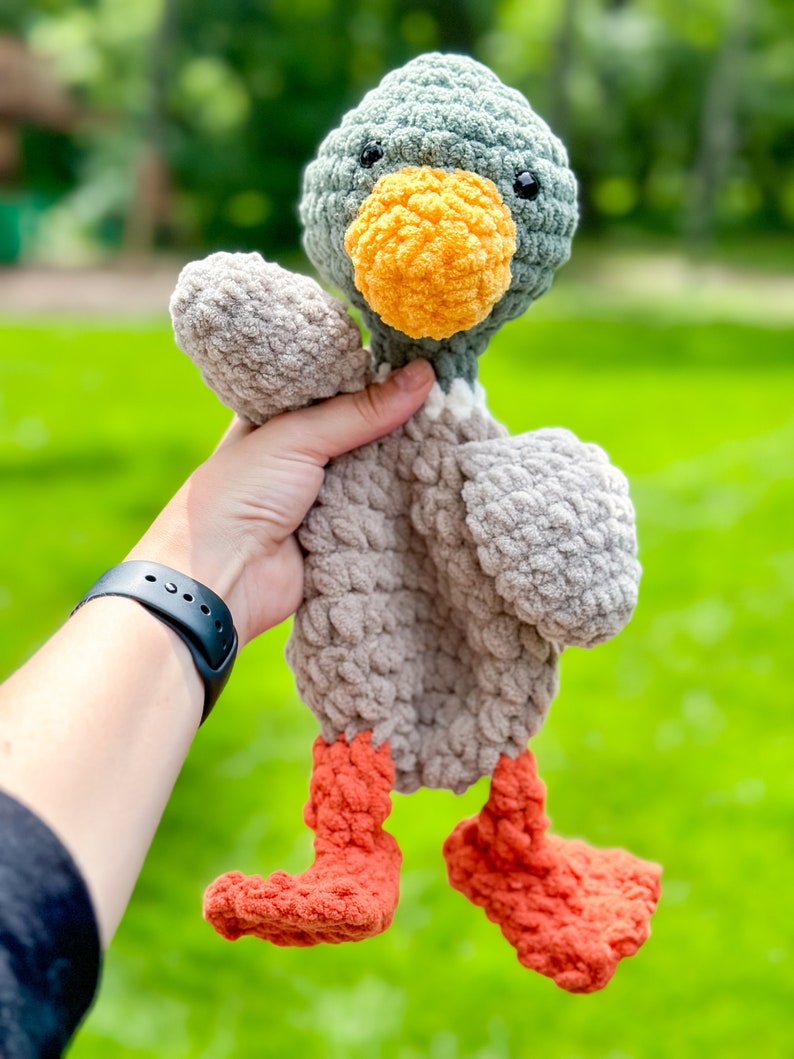Op de afbeelding: Een gehaakt pluche speelgoed eend met een grijs lichaam, een groene kop, een oranje snavel en oranje poten. De eend wordt vastgehouden door een hand.