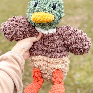 Op de afbeelding: Een pluche eendenspeelgoed met een bruin, beige en oranje lichaam. De eend heeft een groene kop met een gele snavel en zwarte ogen.