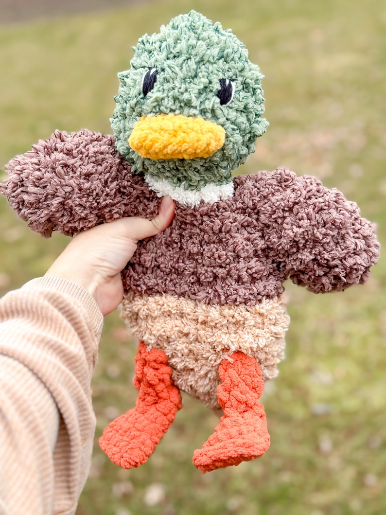 Op de afbeelding: Een pluche eendenspeelgoed met een bruin, beige en oranje lichaam. De eend heeft een groene kop met een gele snavel en zwarte ogen.