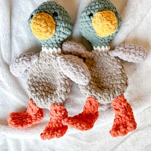 Op de afbeelding: Twee gehaakte pluche speelgoedjes in de vorm van eenden. De eenden zijn grijs met groene koppen en oranje snavels en voeten. Ze liggen op een wit, pluche deken.