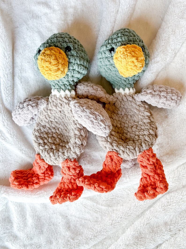 Op de afbeelding: Twee gehaakte pluche speelgoedjes in de vorm van eenden. De eenden zijn grijs met groene koppen en oranje snavels en voeten. Ze liggen op een wit, pluche deken.