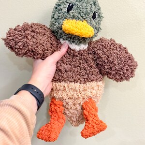 Op de afbeelding: Een pluche eendje met bruin, beige en oranje bont. De eend heeft een groene kop met zwarte ogen en een gele snavel.