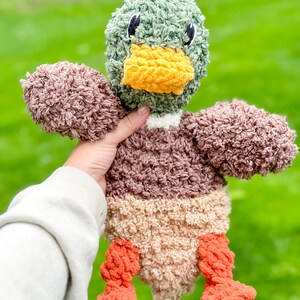 Op de afbeelding: Een pluche eendje met een groene kop, een bruin en beige lichaam en oranje poten. De eend heeft een gele snavel en zwarte ogen.
