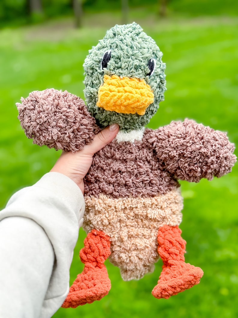 Op de afbeelding: Een pluche eendje met een groene kop, een bruin en beige lichaam en oranje poten. De eend heeft een gele snavel en zwarte ogen.