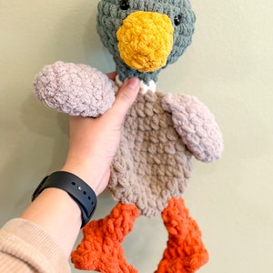 Op de afbeelding: Een gehaakt pluche speelgoed eend met een grijs hoofd, een bruin lichaam en oranje poten. De eend heeft een gele snavel en zwarte ogen.