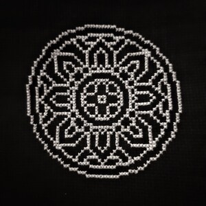 May include: A black and white cross stitch design of a circular mandala pattern. The design is intricate and detailed, with a central cross shape and radiating lines.