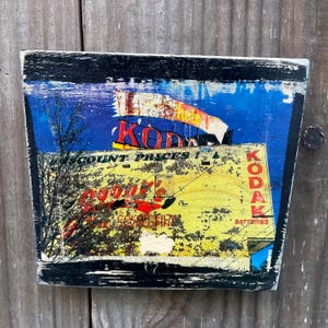 May include: A vintage wooden sign with a faded image of a Kodak store. The sign is painted in yellow, red, and blue with the text "Kodak" and "Discount Prices" visible. The sign has a black border and is slightly weathered.