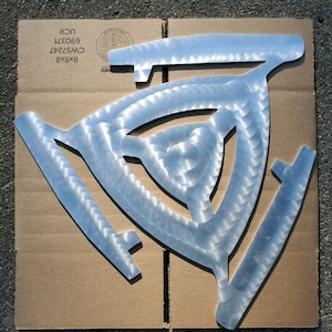 May include: A silver metal camping stove with a triangular design. The stove is sitting on a brown cardboard box with the text "COASTWIDE 8X8 CW57247 690371 UCR" printed on it.