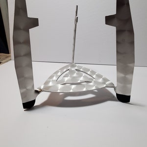 May include: A silver metal stand with a triangular base and three legs. The stand has a textured surface and a small, round opening in the center.
