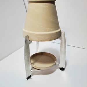 May include: A terracotta pot with a metal stand. The pot is beige and the stand is silver. The stand has three legs and a circular platform.