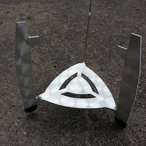 May include: A silver metal tripod stand with a triangular top and three legs. The stand is designed to hold a cooking pot or other cookware.