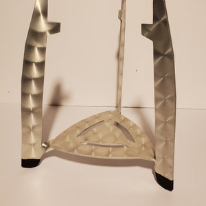 Puede incluir: Un soporte de metal plateado con una base triangular y tres patas. El soporte tiene una superficie texturizada y pies de goma negros.
