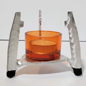May include: A silver metal candle holder with a round, orange glass candle holder on top. The candle holder has a decorative design on the sides.