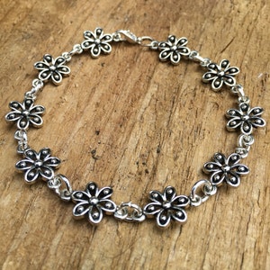 May include: A silver-colored bracelet featuring a daisy flower design. The bracelet is composed of multiple daisy-shaped charms linked together with small chain segments. The bracelet is displayed on a wooden surface.