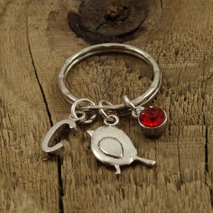 Könnte beinhalten: Silberner Schlüsselanhänger mit einem kleinen Vogel-Anhänger, einem Buchstaben C-Anhänger und einem roten Strassstein. Der Schlüsselanhänger liegt auf einer Holzoberfläche.