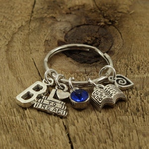 May include: A silver charm keyring with a blue gem, a book charm, a heart charm, and a charm that says "To Read". The keyring is on a wooden surface.