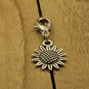May include: Silver sunflower charm with a clip on the top. The charm is on a wooden surface.