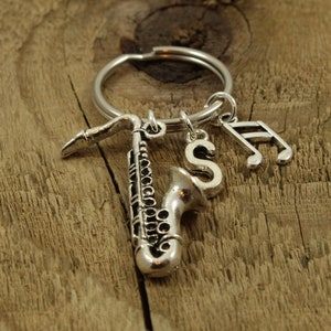 Könnte beinhalten: Silberfarbener Schlüsselanhänger mit einem Saxophon-Anhänger, einer Musiknote und dem Buchstaben "S". Die Anhänger hängen an einem silbernen Ring. Der Hintergrund ist eine rustikale Holzoberfläche.