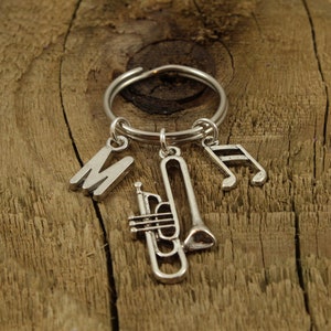Puede incluir: Llavero de plata con un dije de trompeta, un dije de nota musical y un dije de la letra "M". El llavero está sobre una superficie de madera.