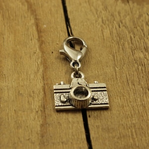 Può includere: Un ciondolo a forma di macchina fotografica color argento con chiusura a moschettone. Il ciondolo della fotocamera presenta un design dettagliato, tra cui un obiettivo, pulsanti e una superficie strutturata. Il ciondolo è sospeso su uno sfondo di legno.