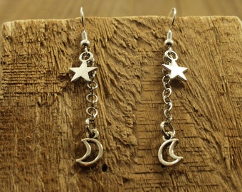 May include: Silver-toned dangle earrings featuring a star and crescent moon charm. The star is at the top, connected by a chain to the moon. The earrings hang from a silver hook. The earrings are displayed on a wooden surface.