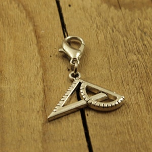 Peut inclure: Charm argenté représentant un triangle rectangle et un rapporteur, attaché à un fermoir. Le bijou est probablement en métal et se détache sur un fond en bois.