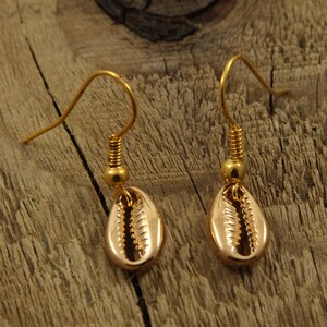 May include: A pair of gold-tone earrings with a seashell design. The earrings are hanging from a wooden surface.