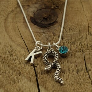 May include: Silver necklace featuring a letter "K" charm, a snake pendant, and a blue gemstone. The necklace is presented on a wooden background.