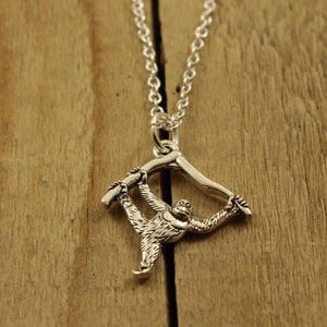 Puede incluir: Collar con colgante plateado con un diseño detallado de orangután. El orangután se representa colgado de una rama, con los brazos extendidos. El collar cuelga de una delicada cadena plateada, sobre un fondo de madera.