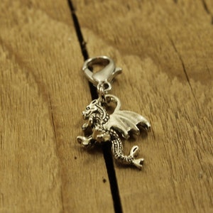 May include: Silver dragon charm with a clip on the top. The dragon is facing right with its wings spread out. The charm is on a wooden surface.