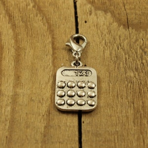 Peut inclure: Charm en forme de calculatrice argentée avec un fermoir mousqueton. La calculatrice a une forme rectangulaire aux bords arrondis, avec une grille de boutons circulaires et les chiffres "123" en haut. Le charme est posé sur un fond en bois.