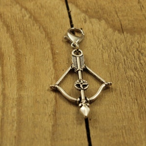 Può includere: Ciondolo d'argento a forma di arco e freccia. La freccia ha un design decorativo e l'arco è aperto.