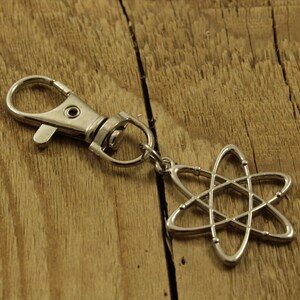 Peut inclure: Porte-clés atome argenté avec mousqueton. Le symbole de l'atome est une représentation stylisée du noyau et des électrons en orbite. Le porte-clés est présenté sur une surface en bois.