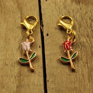 May include: Two rose charms with gold-colored clasps. One charm features a pink rose, the other a red rose, both with green leaves and gold stems. The charms are set against a wooden background.