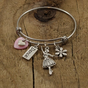 Op de afbeelding: Zilverkleurige armband met bedels. Bedels zijn onder meer een roze hart, een label met het woord "Dance", een ballerina en een bloem. De armband wordt gepresenteerd op een houten oppervlak.
