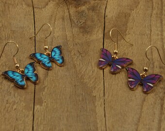 May include: A pair of dangle earrings featuring butterfly designs. One pair is blue and the other is purple.
