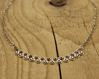 May include: A silver necklace with a delicate chain and a row of small, circular pendants. Each pendant has a cutout design resembling a stylized eye.